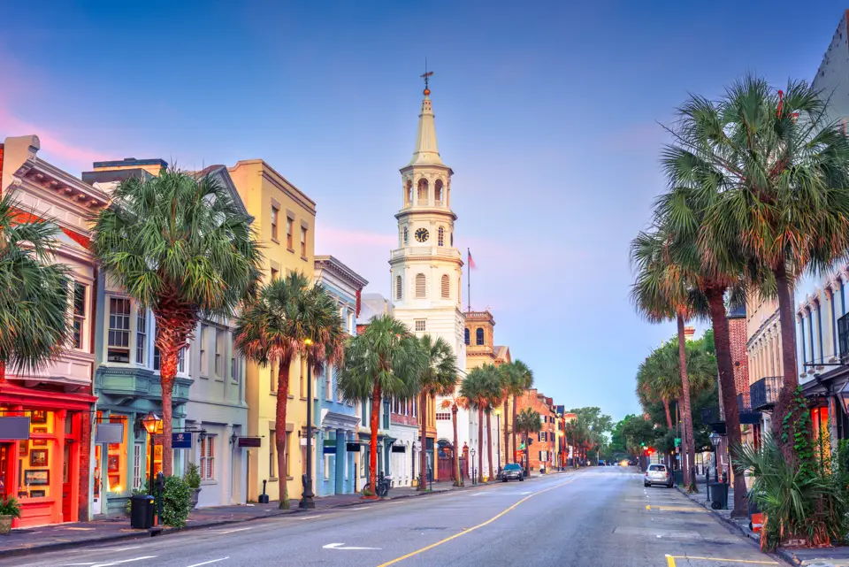 A street lined with palm trees and colorful historic buildings, featuring a prominent clock tower under a clear blue sky.