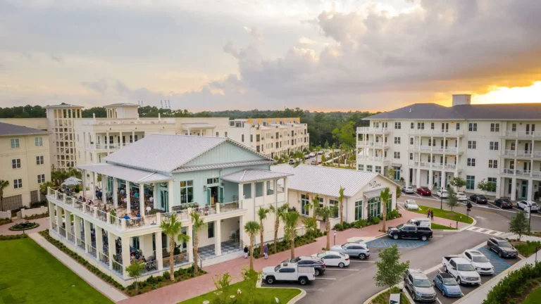 Aerial view of a large residential complex with modern buildings, a central courtyard, parking spaces, and a sunset in the background.