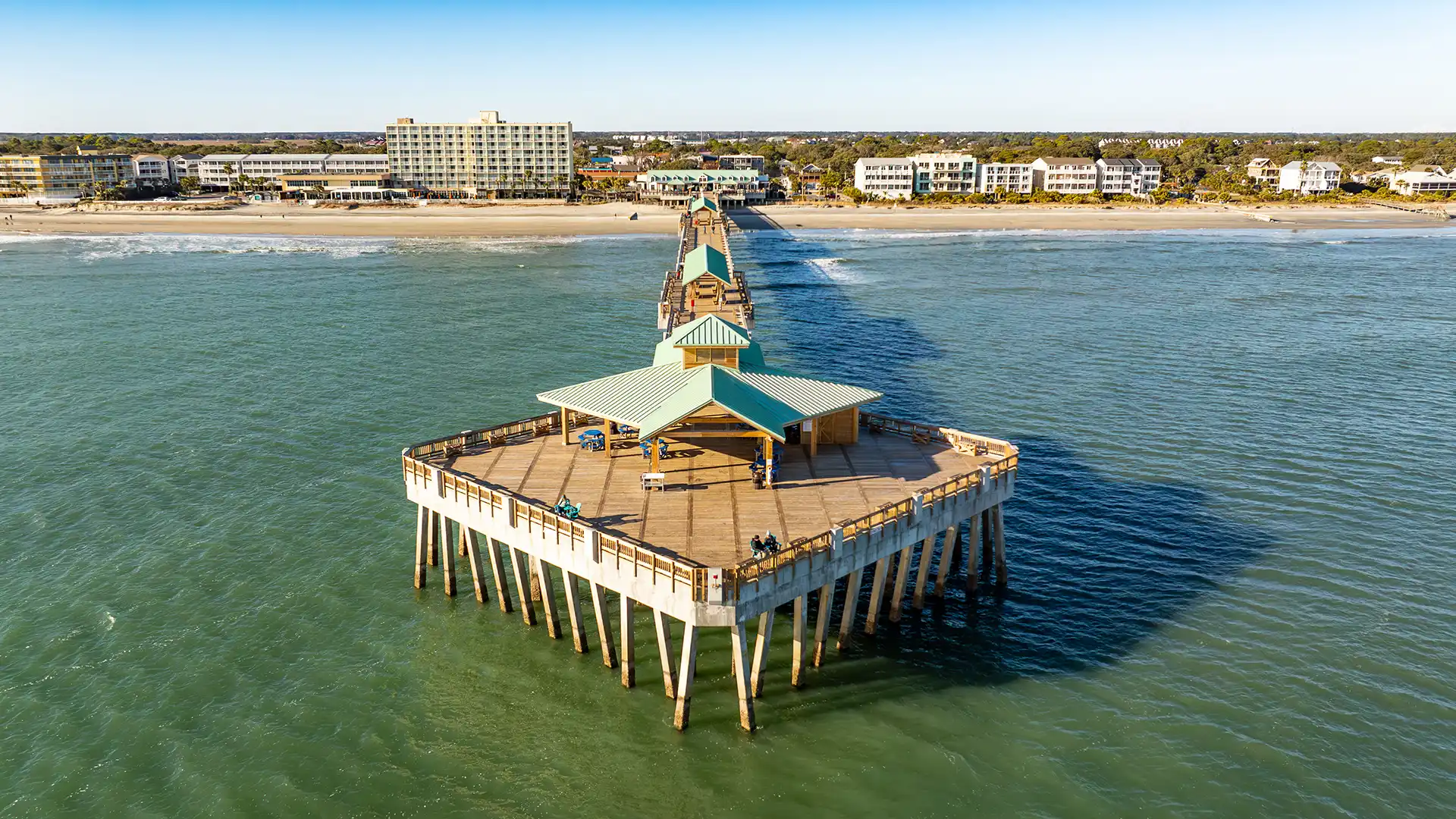 Aerial view of a long wooden pier extending into the ocean with buildings and a sandy beach in the background.