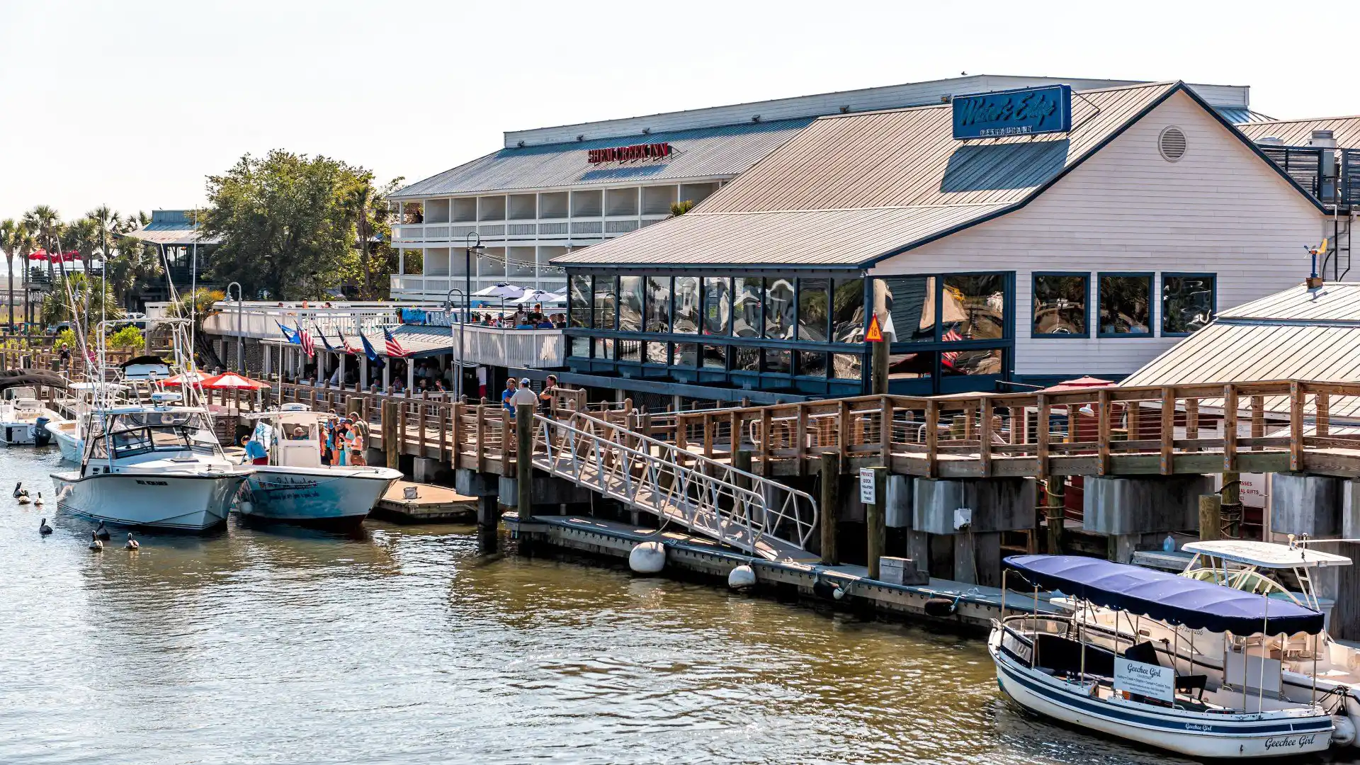 A waterfront restaurant with a wooden dock, several boats moored, and people walking along the pier.