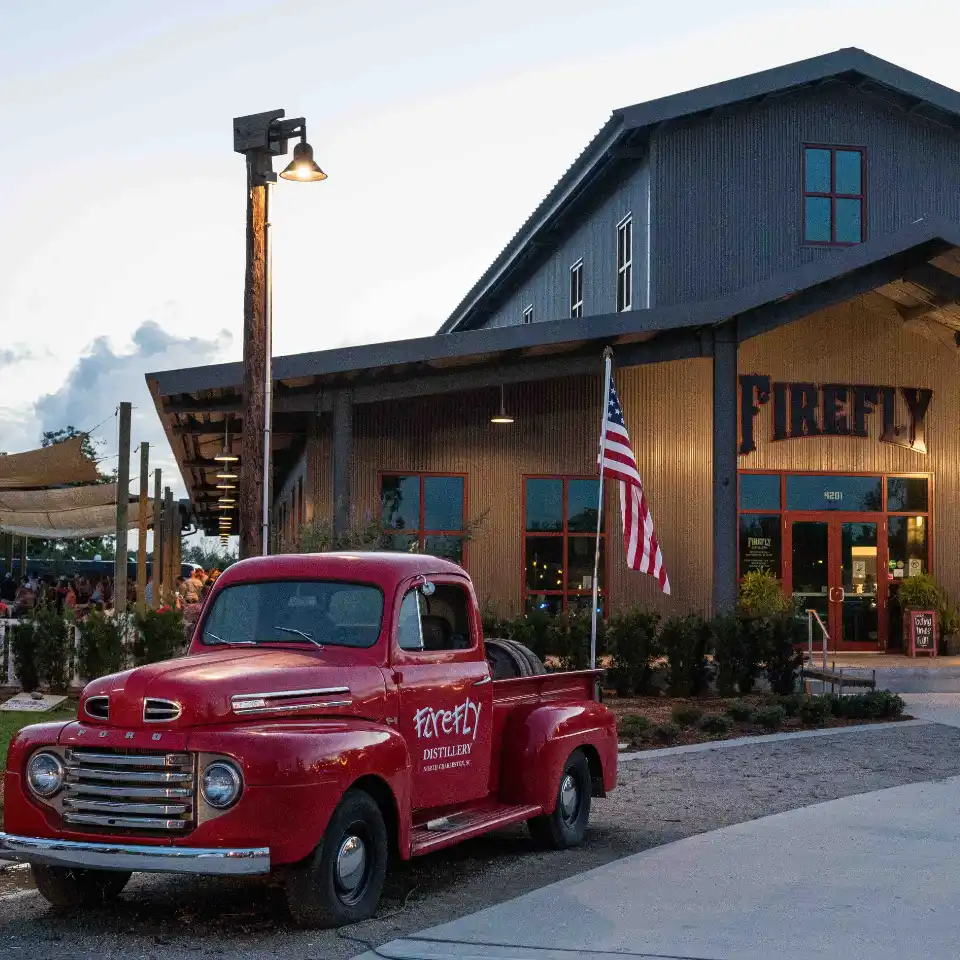 Red vintage truck parked outside Firefly Distillery building at dusk with an American flag.