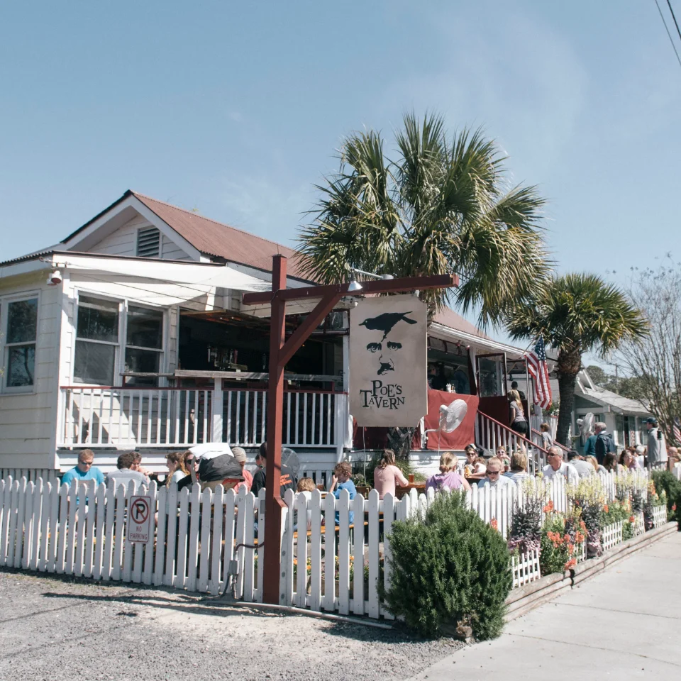 A lively outdoor scene at Poe's Tavern with people seated behind a white picket fence and a backdrop of palm trees.