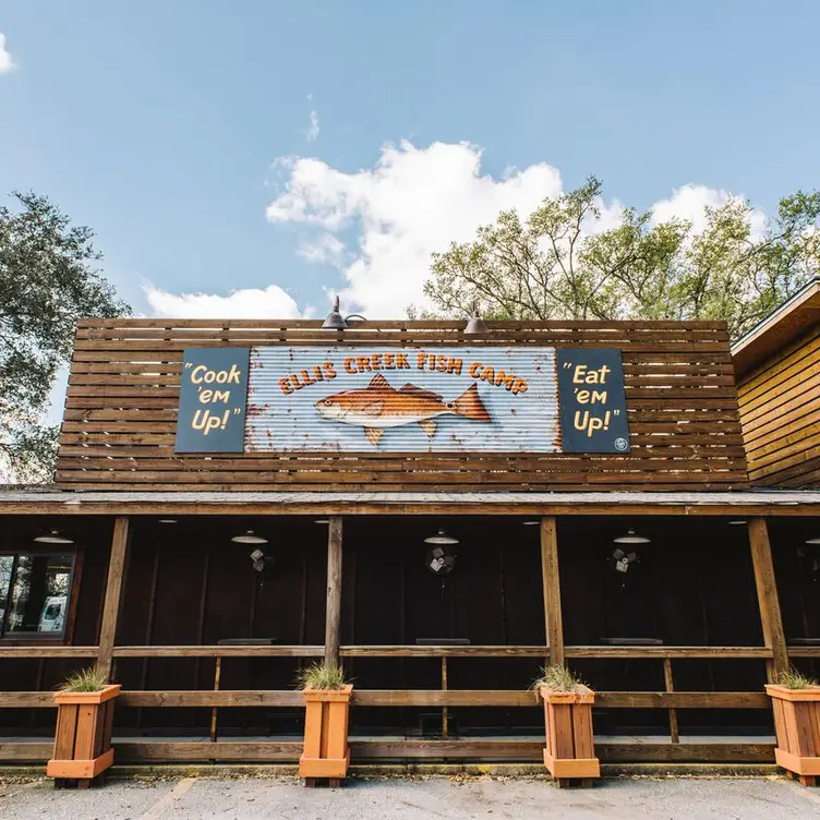 Front view of Ella's Creek Fish Camp restaurant with a large fish sign and slogans "Cook 'em Up!" and "Eat 'em Up!" against a wooden exterior.