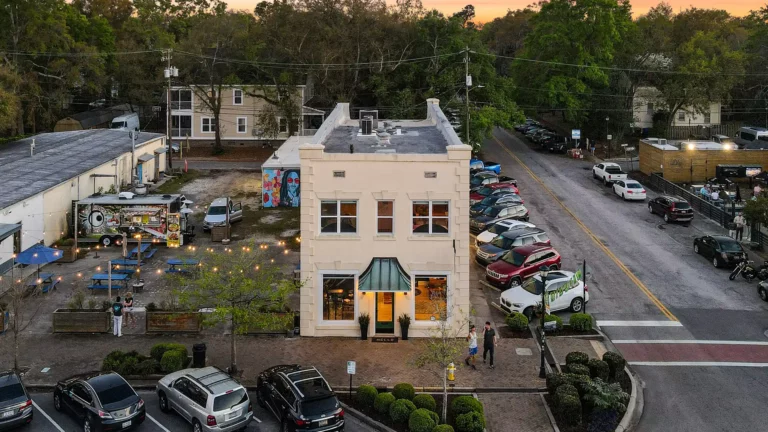 Aerial view of a small two-story building at an intersection, surrounded by parked cars, trees, and nearby businesses with outdoor seating and string lights.