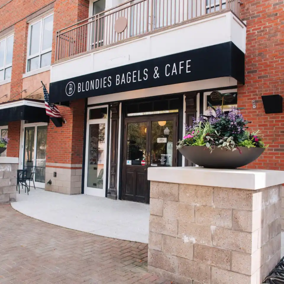 Front entrance of Blondies Bagels & Cafe with a black and white sign, brick exterior, and potted flowers.