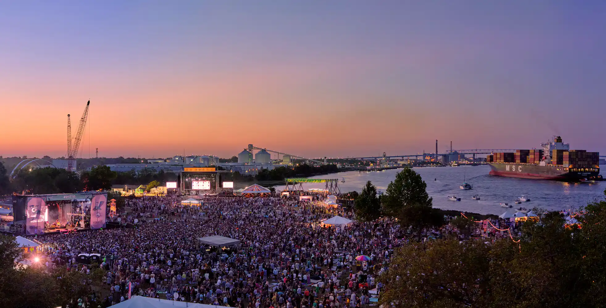 A large crowd gathers at an outdoor festival near a river at sunset, with a cargo ship passing by and stage lights illuminating the event.