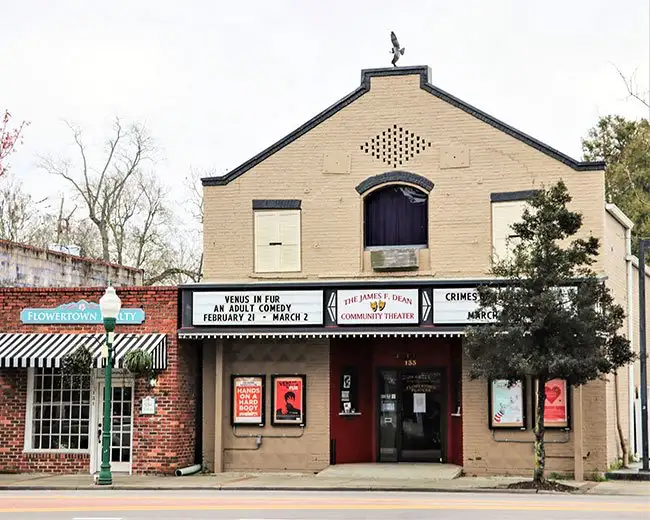 Historic theater building with a marquee advertising upcoming shows, flanked by a tree and a neighboring storefront.